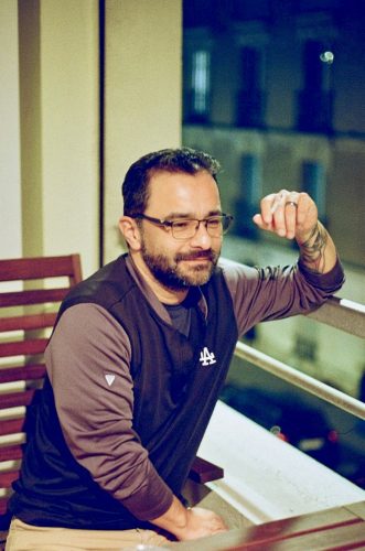 Rudy Martinez, a Chicano writer and podcaster in Paris, relaxes on his balcony at night wearing a Los Angeles Dodgers shirt, enjoying a quiet moment with city lights glowing in the background.